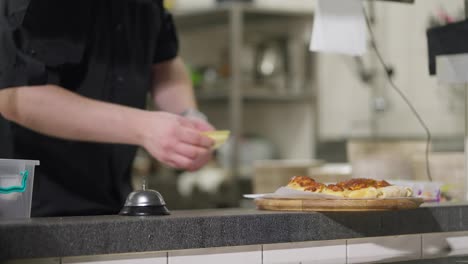 Un-Chef-Profesional-Seguro-De-Sí-Mismo,-Con-Un-Uniforme-Negro,-Coloca-Un-Plato-Terminado-De-Pan-Plano-Con-Queso-Sobre-La-Mesa-Para-Distribuirlo-Y-Hace-Sonar-La-Campana-Para-Señalar-Que-El-Plato-Está-Listo.