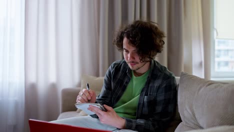 Confident-guy-with-curly-hair-in-a-plaid-shirt-takes-notes-with-a-pen-and-notepad-while-watching-a-video-on-the-couch-at-home