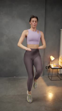 Vertical-video-of-a-confident-brunette-girl-in-a-lilac-top-and-purple-sweatpants-doing-sports-and-running-on-the-spot-at-home-in-a-modern-apartment