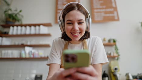 Happy-brunette-girl-in-white-wireless-headphones-T-shirt-and-apron-leans-on-the-counter-and-listens-to-music-in-a-cafe