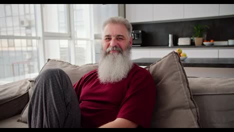 Porträt-Eines-Glücklichen-älteren-Mannes-Mit-Grauem-Haar-Und-üppigem-Bart-In-Einem-Roten-T-Shirt,-Der-Auf-Einem-Sofa-In-Einem-Modernen-Apartment-Sitzt