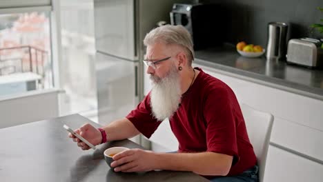 An-elderly-man-with-gray-hair,-a-lush-beard,-glasses-and-a-red-T-shirt-looks-at-his-social-media-feed-and-drinks-tea-during-his-breakfast-in-the-morning-in-a-modern-apartment-overlooking-the-sea