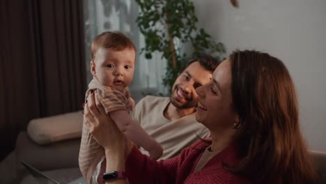 Happy-young-brunette-mother-in-a-red-sweater-together-with-her-brunette-husband-with-her-little-daughter-as-a-baby-and-looking-at-her-in-a-modern-apartment