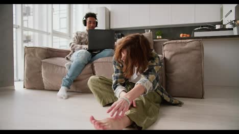 A-young-adult-girl-with-brown-hair-in-a-green-plaid-shirt-sits-on-the-floor-near-the-sofa-and-reaches-for-her-bare-feet-during-a-yoga-class-while-her-young-Black-brunette-boyfriend-works-on-a-gray-laptop-in-a-modern-apartment