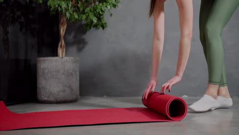 Close-up-a-happy-brunette-girl-in-a-sports-summer-uniform-unrolls-a-red-sports-mat-and-steps-on-it-before-starting-her-sports-activities-in-a-modern-apartment-at-home