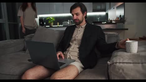 A-brunette-man-with-stubble-in-a-black-jacket-and-beige-shorts-sits-on-the-sofa-and-works-at-a-gray-laptop-drinking-tea-while-his-brunette-girlfriend-hugs-his-shoulders-from-behind-in-a-modern-studio-apartment-in-the-evening