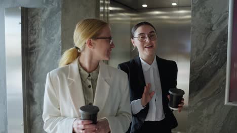 Confident-blonde-girl-in-a-white-business-suit-walks-with-her-brunette-female-colleague-during-the-start-of-their-working-day-and-coffee-break-in-a-modern-office