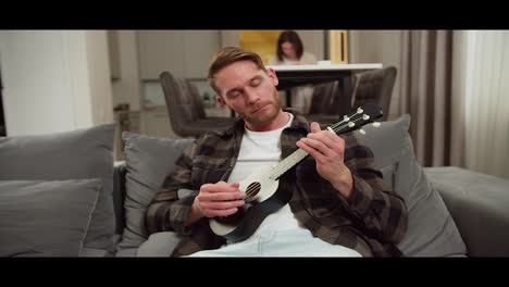 Confident-and-calm-blond-man-with-stubble-in-a-checkered-brown-shirt-plays-on-a-black-musical-string-instrument-ukulele-while-sitting-on-a-gray-sofa-while-his-wife-works-at-the-table-in-a-modern-apartment-during-the-day