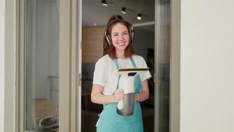 Portrait-of-a-happy-brunette-cleaning-lady-girl-in-black-wireless-headphones-and-a-white-T-shirt-and-a-blue-apron-who-poses-with-a-window-vacuum-cleaner-in-her-hands-near-a-glass-door-in-a-modern-apartment-during-cleaning