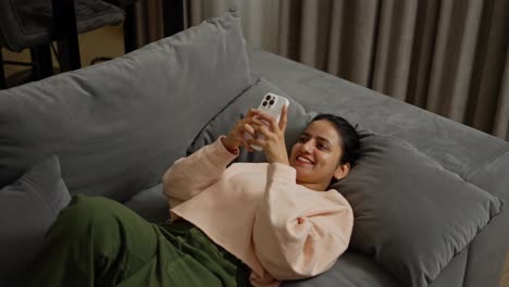 From-above,-a-happy-brunette-girl-in-a-beige-sweater-and-green-pants-lies-on-a-pillow-on-a-gray-sofa-and-actively-scrolls-social-networks-on-her-white-smartphone-during-the-day