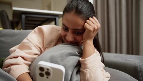 Close-up-a-happy-brunette-girl-with-red-lipstick-in-a-light-jacket-hugs-a-gray-pillow-lying-on-the-sofa-and-types-on-her-smartphone-during-the-day