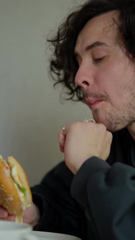 Vertical-video-close-up-of-a-sleepy-brunette-guy-with-a-mustache-and-curly-hair-eating-his-sandwich-in-the-morning-and-enjoying-it