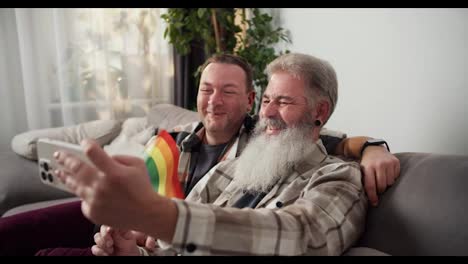 Happy-elderly-man-with-gray-hair-color-and-lush-white-beard-in-a-checkered-shirt-holds-an-LGBT-flag-and-takes-a-selfie-using-a-White-phone-along-with-his-brunette-boyfriend-on-a-gray-sofa-in-a-modern-apartment