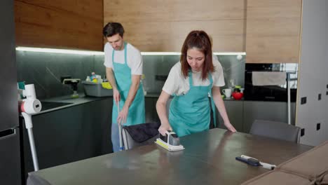 Confident-brunette-girl-in-a-blue-apron-cleaner-polishes-the-surface-of-the-table-with-her-colleagues-while-cleaning-in-a-modern-apartment-in-the-kitchen