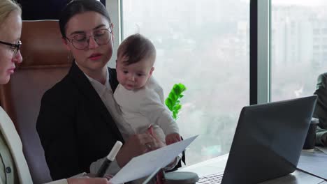 Meeting-of-two-colleagues-Business-Woman-brunette-girl-in-round-glasses-and-a-business-suit-holds-her-small-infant-child-while-communicating-on-serious-issues-with-her-colleagues-at-the-office-table