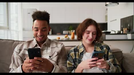 A-happy-man-with-Black-skin-a-brunette-with-stubble-and-his-girlfriend-in-checkered-shirts-are-sitting-on-a-gray-sofa-and-looking-at-programs-in-their-smartphones-and-Social-Media-sitting-on-the-sofa-in-a-modern-apartment