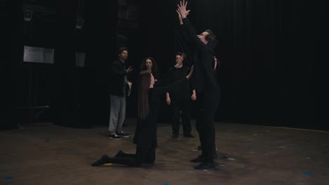 Un-Grupo-De-Actores-Confiados-Con-Trajes-Negros,-Jóvenes-Estudiantes-Universitarios-De-Teatro,-Están-Ensayando-Mostrando-Expresión-Durante-Las-Acciones-Junto-Con-Su-Joven-Director-De-Escena-Masculino-Con-Papeles-En-Sus-Manos-En-El-Escenario.