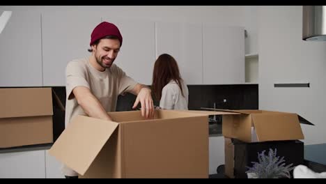 A-happy-man-with-stubble-in-a-red-cap-and-beige-T-shirt-gives-his-brunette-girlfriend-in-a-white-T-shirt-things-from-a-box-after-moving-into-a-new-apartment.-Happy-couple-unpacking-things-from-boxes-after-moving