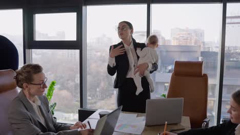 A-confident-brunette-girl-in-round-glasses-in-a-black-business-suit-holds-a-small-infant-child-in-her-hands-and-holds-a-businesswoman-meeting-in-a-modern-office-at-the-table