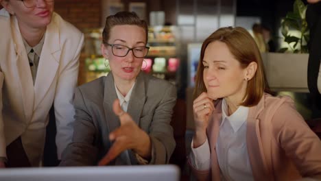 Confident-middle-aged-blonde-girl-with-glasses-in-a-gray-business-uniform-presents-her-work-on-a-laptop-during-a-meeting-of-colleagues-in-a-modern-office