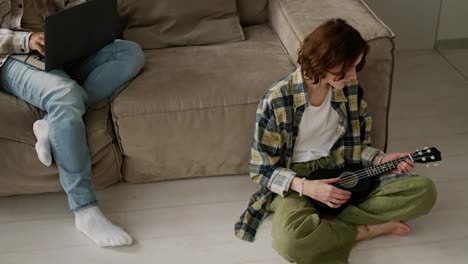 A-girl-with-brown-hair-with-a-bob-hairstyle-in-a-checkered-shirt-plays-the-ukulele-while-her-boyfriend-a-young-brunette-with-Black-skin-color-in-black-headphones-works-on-his-gray-laptop-Sitting-on-the-sofa-in-a-modern-studio-apartment