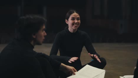 Una-Chica-Morena-Feliz-Con-Un-Traje-Negro,-Una-Actriz,-Se-Sienta-En-El-Escenario-Y-Se-Comunica-Con-Sus-Colegas-Durante-Un-Ensayo-Y-Preparación-Para-Una-Actuación-En-El-Teatro.