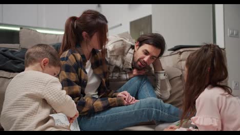 Un-Hombre-Moreno-Feliz-Con-Barba-Incipiente-Que-Viste-Una-Camisa-Beige-Se-Comunica-Con-Su-Pequeña-Hija-Y-Su-Hijo-Junto-Con-Su-Esposa-Mientras-Los-Niños-Dibujan-En-El-Piso-Cerca-Del-Sofá-En-Un-Apartamento-Moderno