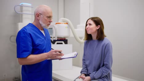 A-confident-elderly-male-doctor-in-glasses-with-a-blue-uniform-communicates-with-a-young-brunette-girl-and-gives-her-advice-on-disease-prevention-during-an-appointment-with-a-doctor-in-a-modern-clinic