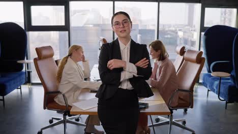 Portrait-of-a-confident-brunette-businesswoman-in-round-glasses-in-a-black-business-suit-who-folds-her-arms-on-her-chest-and-poses-confidently-in-a-modern-office-with-large-windows