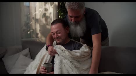 A-happy-elderly-man-with-gray-hair-and-a-lush-beard-wraps-his-brunette-boyfriend-in-a-white-woolen-blanket-while-he-drinks-tea-and-sits-on-the-sofa-in-a-modern-apartment-in-the-evening.-Showing-love-and-care-in-a-happy-LGBT-couple-of-two-men