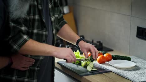 Close-up-a-middle-aged-brunette-man-in-a-plaid-shirt-prepares-a-vegetable-salad-while-his-elderly-boyfriend-with-gray-hair-and-a-bushy-beard-hugs-the-man-and-supports-him-in-the-kitchen-before-breakfast