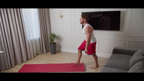 A-happy-blond-man-with-a-beard-and-an-athletic-build-in-a-white-T-shirt-and-red-pants-lays-out-a-red-carpet-and-begins-his-warm-up-before-playing-sports-at-home