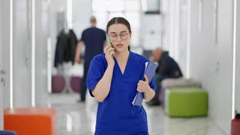 Eine-Selbstbewusste-Brünette-Ärztin-Mit-Runder-Brille-Und-Blauer-Uniform-Geht-Den-Flur-Entlang-Und-Telefoniert-In-Einer-Modernen-Klinik.-Selbstbewusstes-Brünettes-Mädchen-Mit-Runder-Brille,-Arzt-Kommuniziert,-Geht-In-Einer-Modernen-Klinik-Auf-Abruf