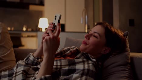 A-happy-and-confident-brunette-girl-in-a-plaid-shirt-lies-on-a-gray-sofa-and-types-on-social-networks-using-her-white-smartphone-in-the-evening-in-a-modern-apartment