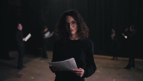 Portrait-of-a-confident-brunette-girl-with-curly-hair-in-glasses-and-a-black-actors-suit-with-papers-in-her-hands-in-a-group-of-actors-who-are-rehearsing-before-performing-on-stage-in-a-theater-with-black-curtains