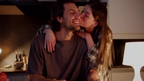 Happy-brunette-girl-in-an-orange-T-shirt-and-plaid-shirt-approaches-her-boyfriend-from-behind-and-hugs-him.-Happy-brunette-guy-reading-a-book-and-hugging-his-brunette-girlfriend-in-a-modern-apartment-in-the-kitchen-in-the-evening