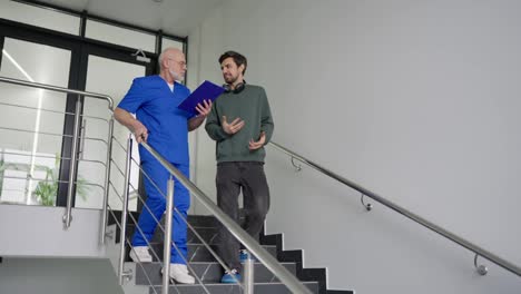 Un-Joven-Moreno-Con-Una-Chaqueta-Verde-Y-Auriculares-Inalámbricos-Se-Comunica-Con-Un-Médico-Experimentado-Con-Barba-Gris-Y-Uniforme-Azul-Que-Escribe-Las-Quejas-De-Los-Pacientes-En-Su-Tableta-Mientras-Baja-Las-Escaleras-En-Una-Clínica-Moderna.