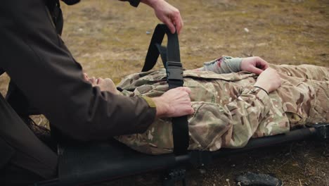 Close-up-a-confident-man-military-man-in-dark-green-army-clothes-fastens-a-wounded-soldier-in-camouflage-clothing-on-a-stretcher-with-a-belt-in-combat-conditions-in-the-steppe