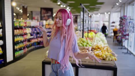 Niña-Feliz-Con-Cabello-Rosado-Que-Usa-Auriculares-Inalámbricos-Escuchando-Música-Y-Bailando-En-Una-Tienda-De-Comestibles