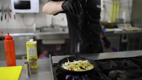 Close-up-of-a-confident-man-Chef-in-a-black-uniform-sprinkles-vegetables-with-salt-which-he-fries-in-a-frying-pan-on-the-stove-in-a-restaurant-kitchen