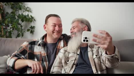 Feliz-Pareja-Gay:-Un-Hombre-De-Mediana-Edad-Con-Cabello-Gris-Y-Una-Barba-Exuberante-Se-Toma-Una-Selfie-Usando-Un-Teléfono-Blanco-Junto-Con-Su-Novio-Moreno-Con-Una-Camisa-A-Cuadros-Mientras-Está-Sentado-En-El-Sofá-De-Un-Apartamento-Moderno