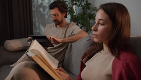 Close-up-view-of-a-young-brunette-girl-in-a-red-sweater-reading-a-book-while-her-brunette-boyfriend-with-stubble-turns-his-attention-to-her-on-the-sofa-in-a-modern-apartment-at-home