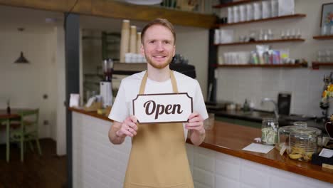 Porträt-Eines-Glücklichen-Brünetten-Kellners-In-Einer-Gelben-Schürze,-Der-In-Einem-Café-Offen-Ein-Schild-In-Den-Händen-Hält