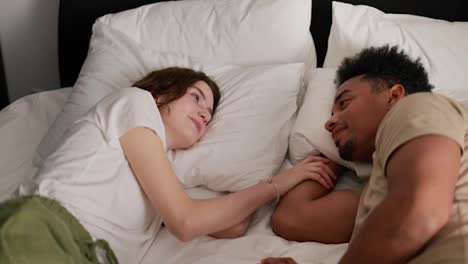 A-happy-young-adult-brunette-girl-with-a-bob-hairstyle-in-a-white-T-shirt-lies-on-the-bed-with-her-young-boyfriend-with-Black-brunette-skin-and-touches-his-hand-in-the-morning