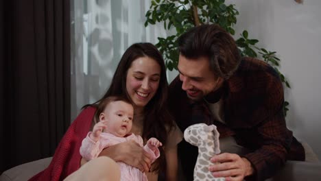 Happy-brunette-man-in-a-brown-shirt-together-with-his-young-brunette-wife-plays-with-his-little-baby-daughter-in-a-pink-overalls-while-sitting-on-a-gray-sofa-in-a-modern-apartment