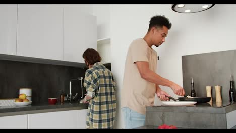 A-young-man-with-Black-skin-color-in-a-beige-T-shirt-puts-ready-made-scrambled-eggs-on-a-plate-while-his-young-adult-girlfriend-with-a-bob-hairstyle-washes-the-dishes-while-preparing-breakfast-in-the-morning-at-a-modern-apartment