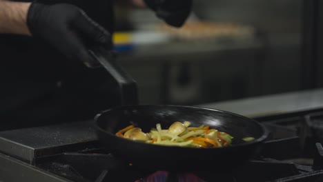 Close-up-a-professional-chef-in-a-black-uniform-mixes-thinly-sliced-vegetables-on-a-hot-frying-pan-while-preparing-a-dish-on-a-gas-burner-in-a-restaurant-kitchen