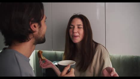 A-happy-brunette-girl-in-a-beige-T-shirt-is-trying-to-get-a-piece-of-bun-stuck-in-her-teeth-and-her-brunette-boyfriend-with-stubble-helps-her-and-they-are-having-fun-together-while-spending-time-together-in-the-evening-in-a-modern-kitchen