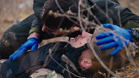 Primer-Plano-De-Un-Hombre-Moreno-Con-Barba-Con-Uniforme-Verde-Oscuro-Y-Guantes-Médicos-Azules-Escucha-La-Respiración-De-Un-Soldado-Herido-Con-Uniforme-De-Camuflaje-Y-Chaleco-Antibalas-Que-Yace-En-El-Suelo-En-La-Estepa-Durante-Operaciones-De-Combate