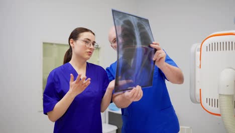 Un-Médico-Experimentado,-Un-Anciano-Con-Gafas-Y-Uniforme-Azul,-Se-Comunica-Y-Consulta-Con-Una-Joven-Morena,-Un-Médico-Con-Gafas,-Mientras-Realiza-Los-Resultados-De-La-Fluorografía-En-Una-Clínica-Moderna.